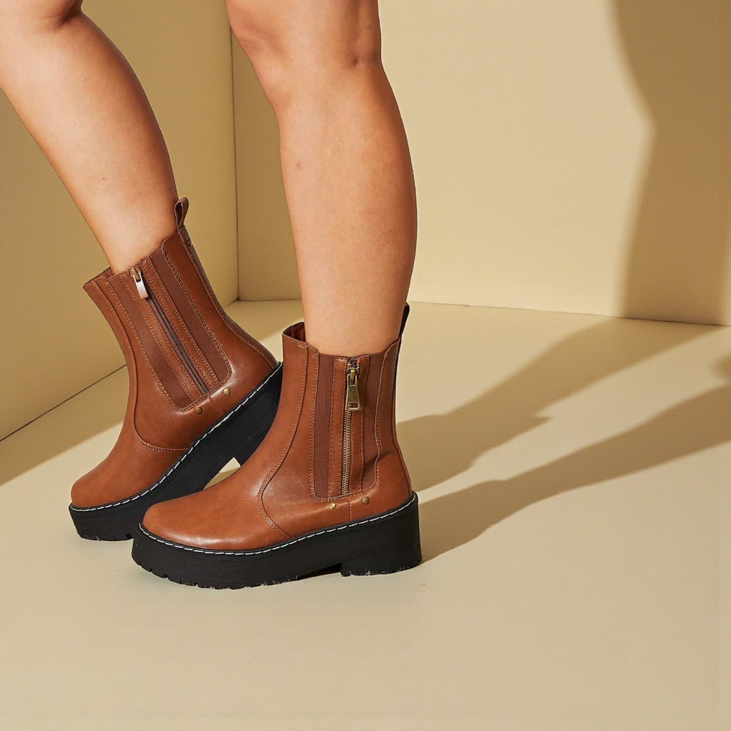 Brown ankle boots with black soles worn by a person on a beige background