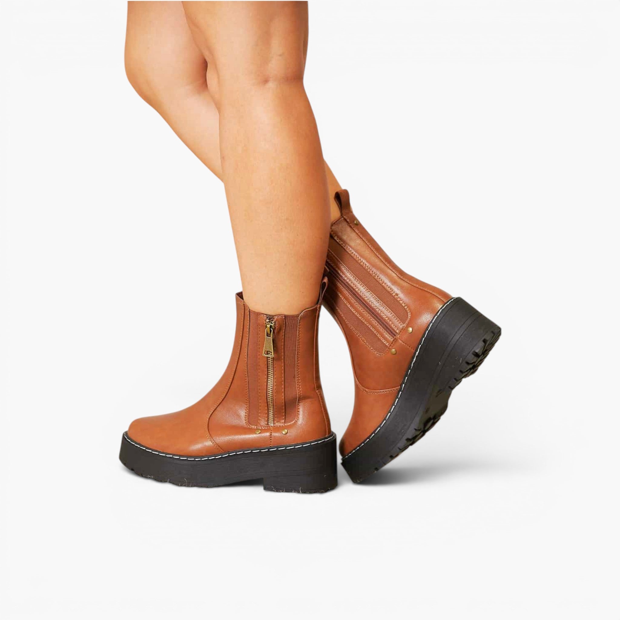 A pair of women's ankle boots in tan brown leather with black platform soles and side zippers, shown on a model's feet against a light gray studio background.
