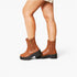 A pair of women's brown leather ankle boots with black platform soles and side zippers, shown on a model's feet against a light gray studio background.