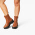 A pair of women's ankle boots in tan brown leather with black platform soles and side zippers, shown on a model's feet against a light gray studio background.