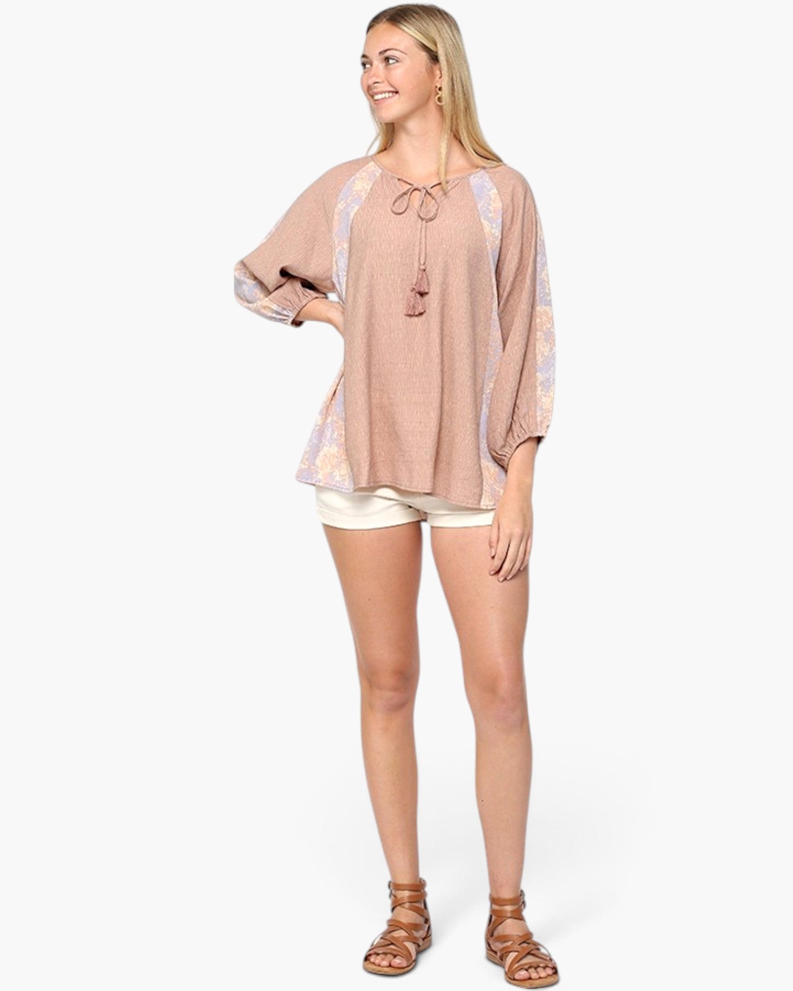 A woman modeling a loose, dusty pink bohemian-style top with lavender and cream patterned sleeves, paired with cream shorts and brown strappy sandals, against a light gray studio background.