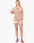 A woman modeling a loose, dusty pink bohemian-style top with lavender and cream patterned sleeves, paired with cream shorts and brown strappy sandals, against a light gray studio background.