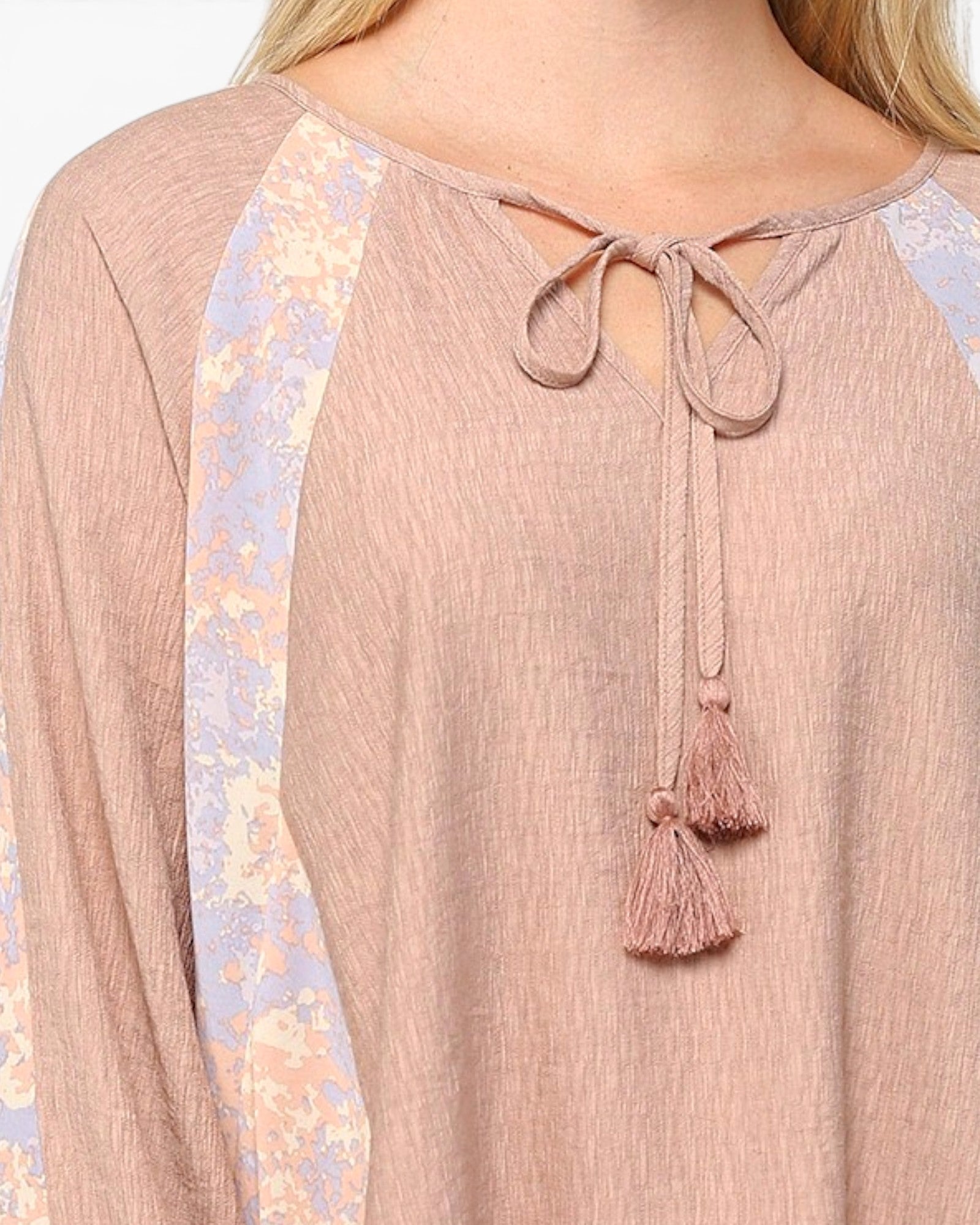 A close-up of a woman wearing a dusty rose long-sleeve top with a tie-neck design and tassels, featuring light lavender and cream floral patterned raglan sleeves.