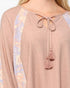 A close-up of a woman wearing a dusty rose long-sleeve top with a tie-neck design and tassels, featuring light lavender and cream floral patterned raglan sleeves.