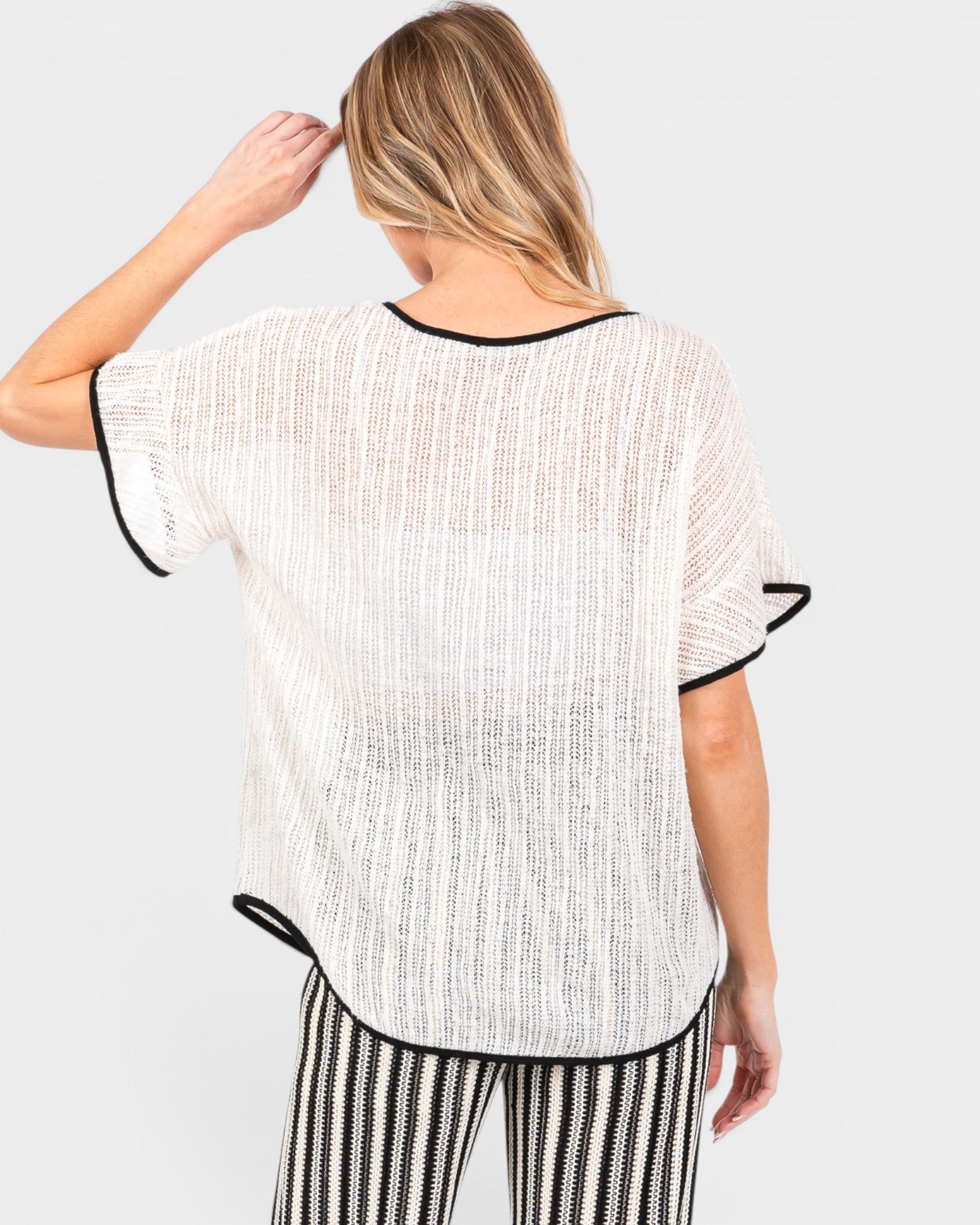 A woman shown from the back wearing a loose white textured short-sleeve top with black trim, paired with black-and-white striped pants, against a light gray studio background.