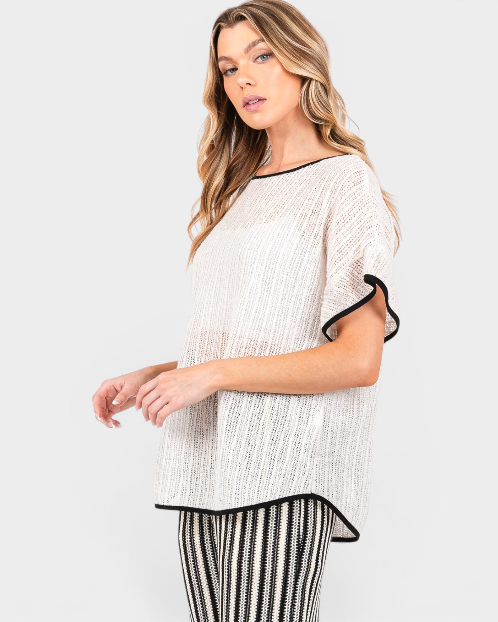 A woman modeling a loose, short-sleeve textured knit top in off-white with black trim, paired with black-and-white striped pants, against a light gray studio background.
