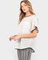 A woman modeling a loose, short-sleeve textured knit top in off-white with black trim, paired with black-and-white striped pants, against a light gray studio background.
