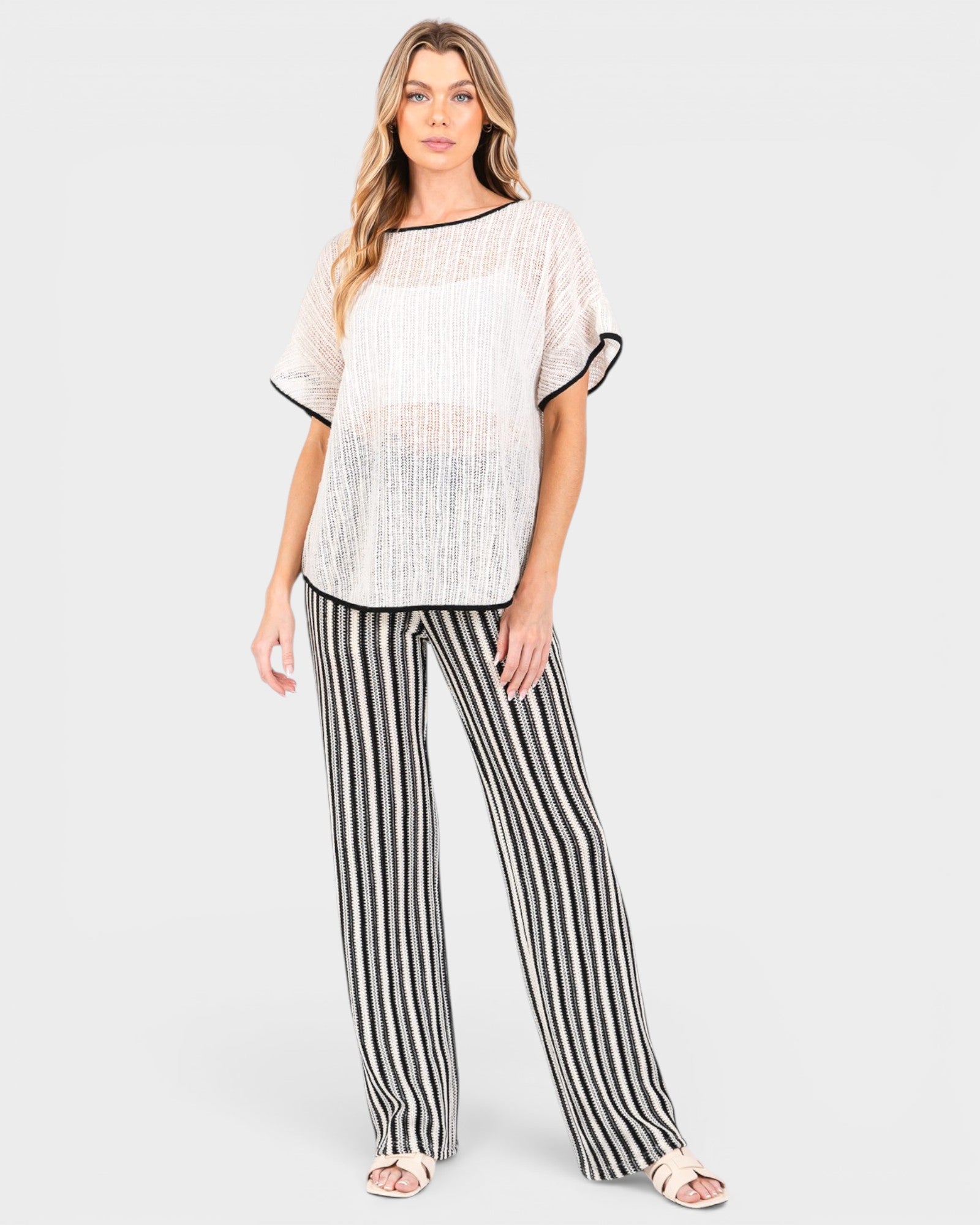 A woman modeling a two-piece lounge set consisting of a white textured short-sleeve top with black trim and black-and-white vertically striped wide-leg pants, set against a plain light gray background.