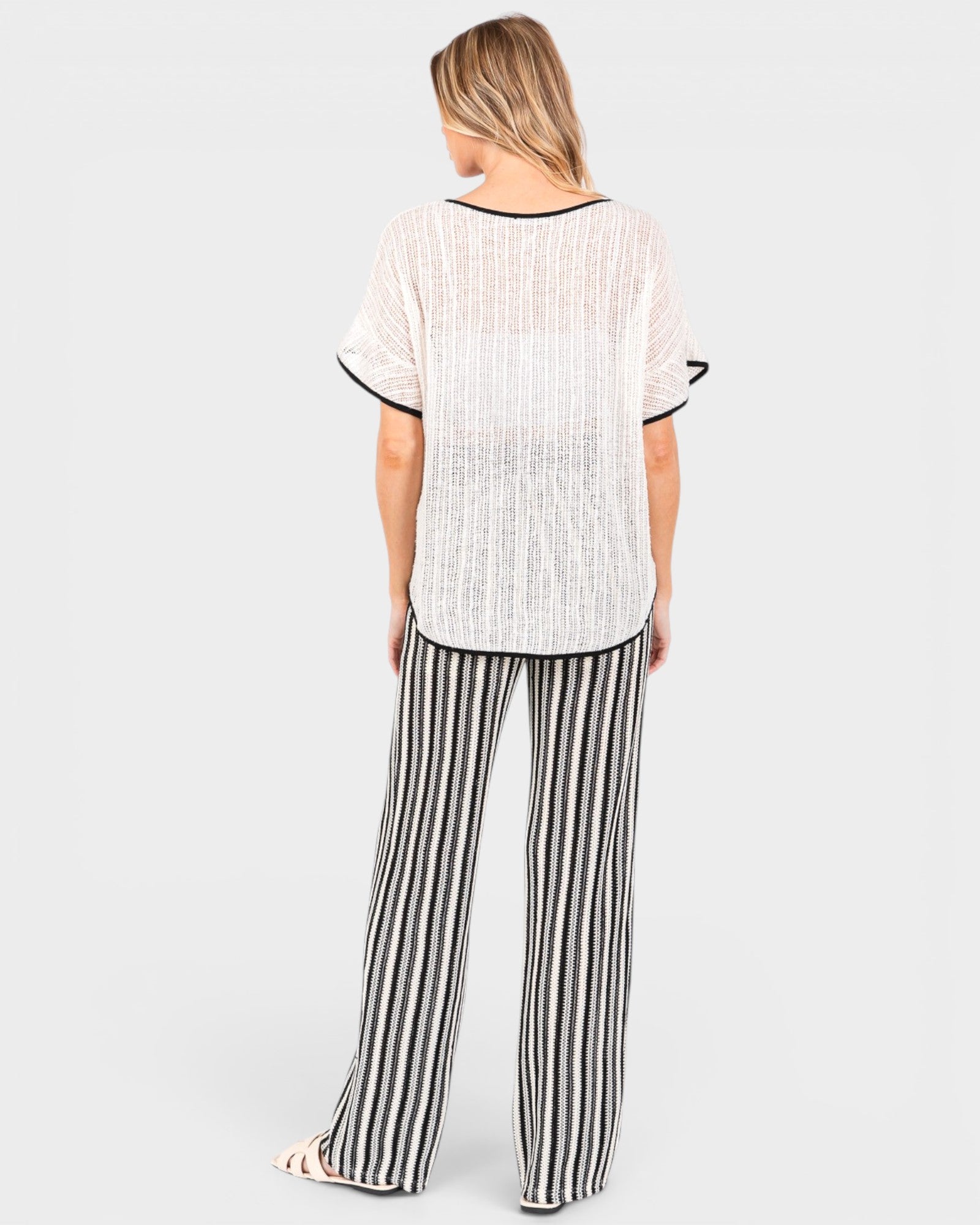 A woman shown from the back wearing a two-piece lounge set consisting of a white textured short-sleeve top with black trim and black-and-white vertically striped wide-leg pants, set against a solid light gray background.