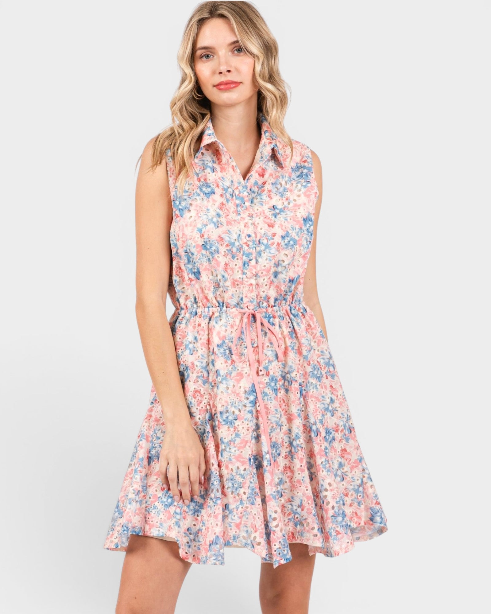 A woman modeling a sleeveless, high-neck, knee-length A-line dress with a pink, blue, and white floral pattern against a light gray studio background.