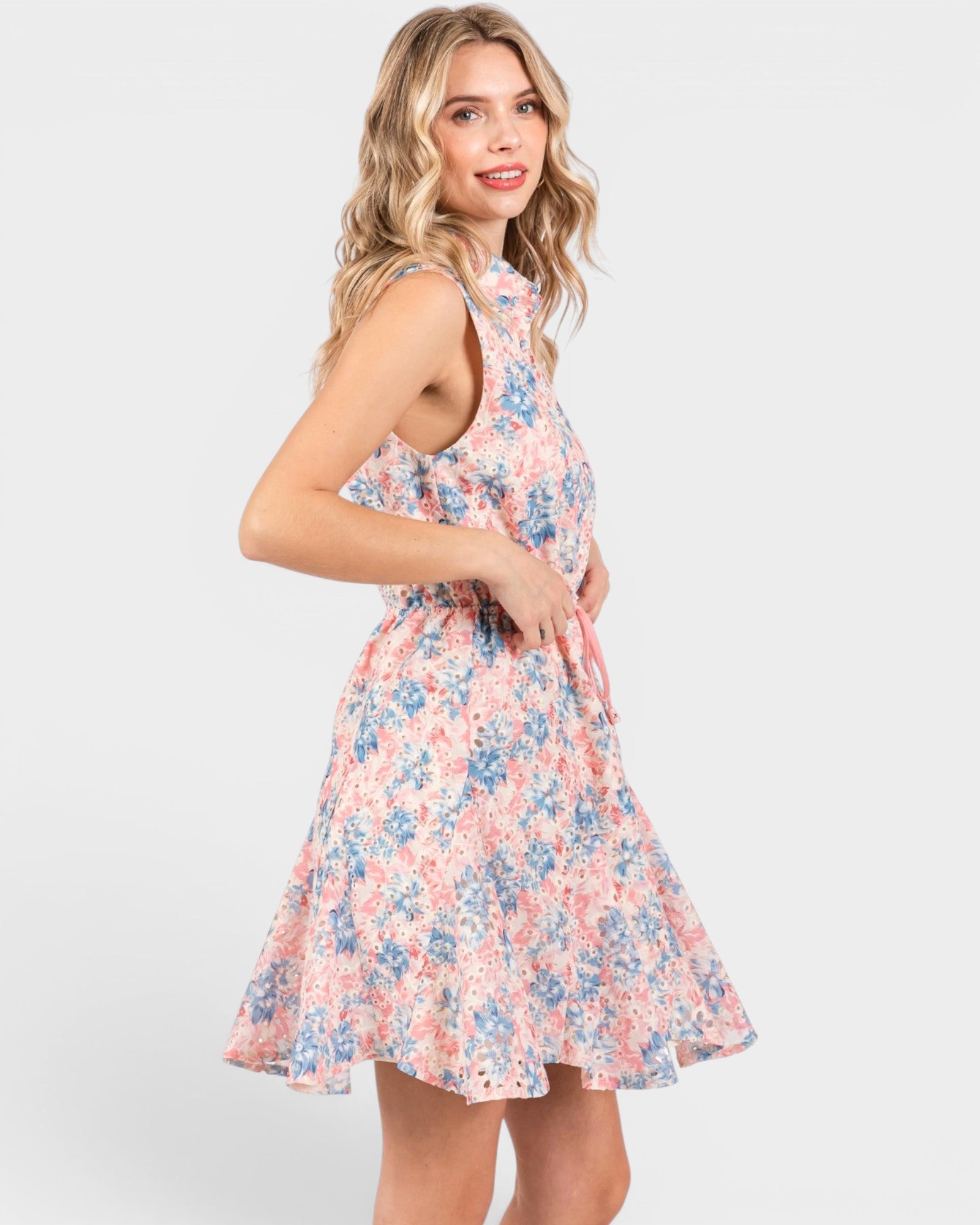 A woman modeling a sleeveless, high-neck, knee-length A-line dress with a pink, blue, and white floral pattern against a light gray studio background.
