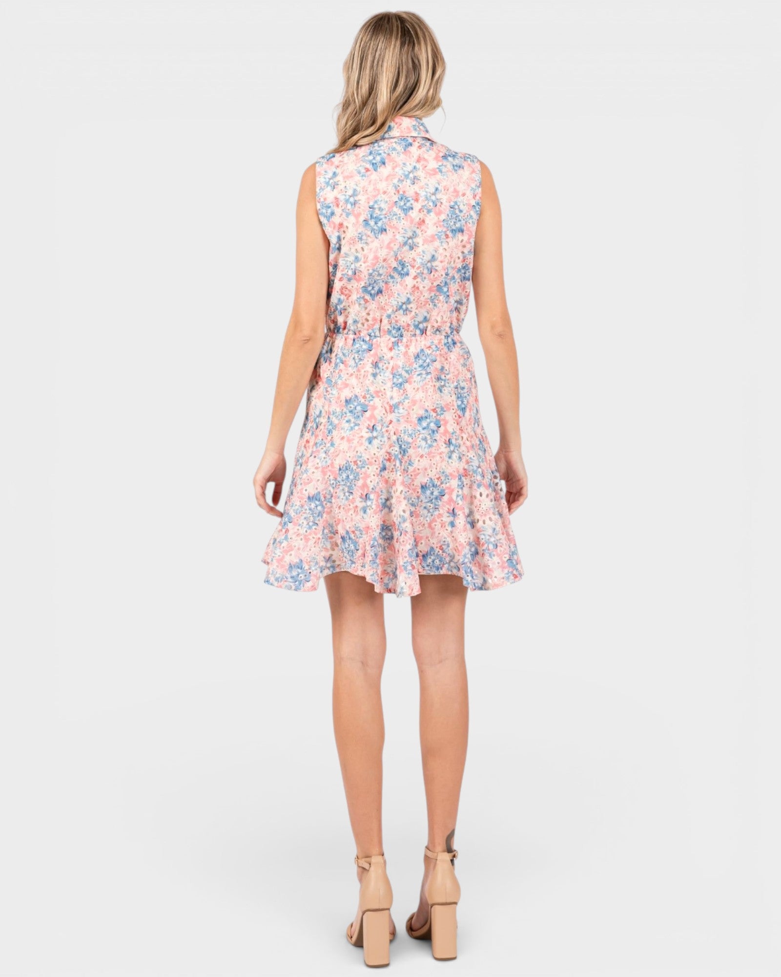 A woman viewed from the back wearing a sleeveless, knee-length floral dress with a ruffled hem and tan high-heeled sandals, against a light gray studio background.
