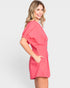 A woman modeling a short pink wrap-style romper with puff sleeves and side pockets, photographed against a light gray studio background.