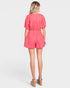A woman viewed from behind wearing a bright coral pink short-sleeve romper with puff sleeves and an elastic waist, paired with tan wedge sandals, against a light gray studio background.