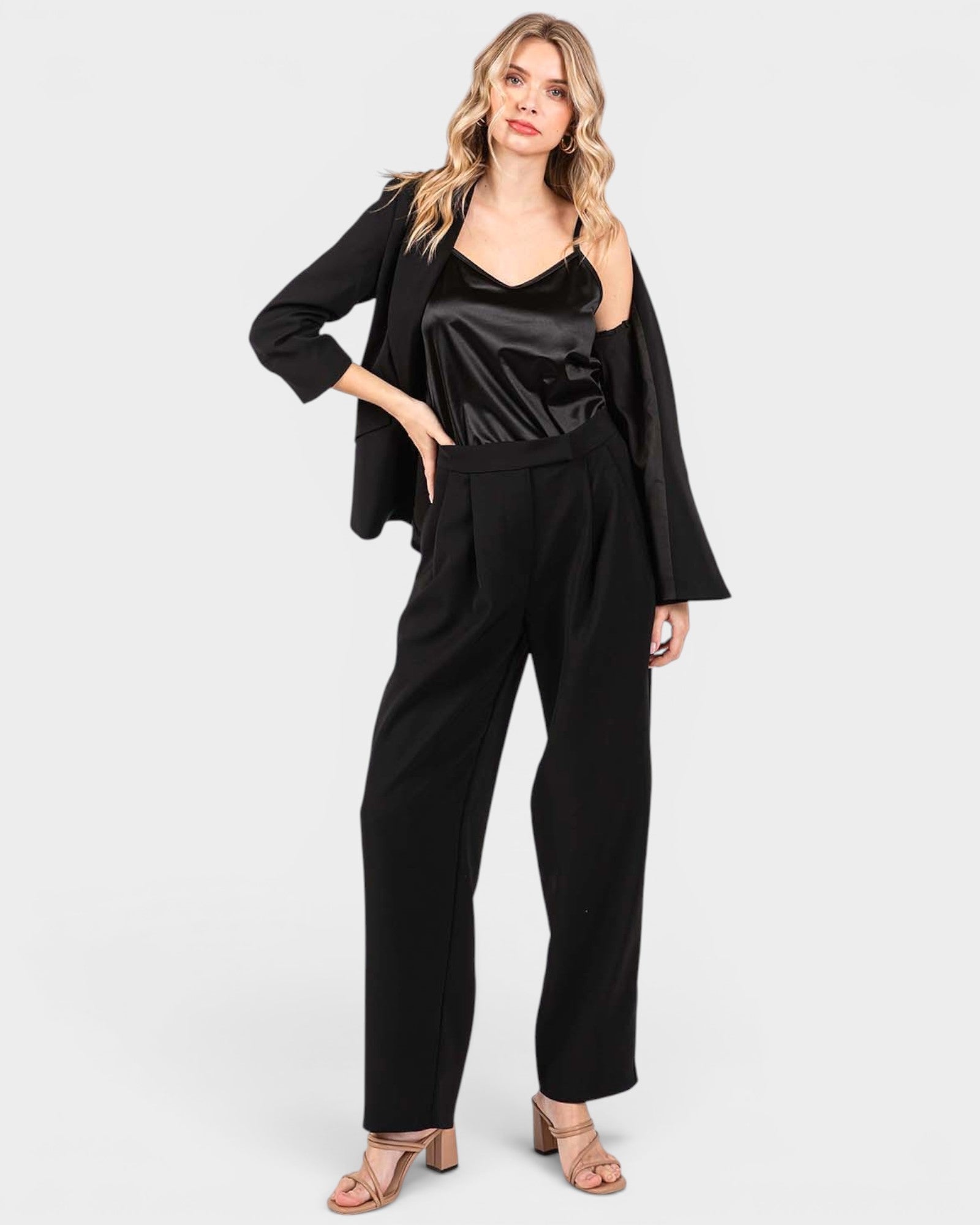 A woman modeling a black three-piece outfit consisting of a satin camisole top, wide-leg trousers, and a draped open-front blazer, paired with beige heeled sandals against a light gray background.