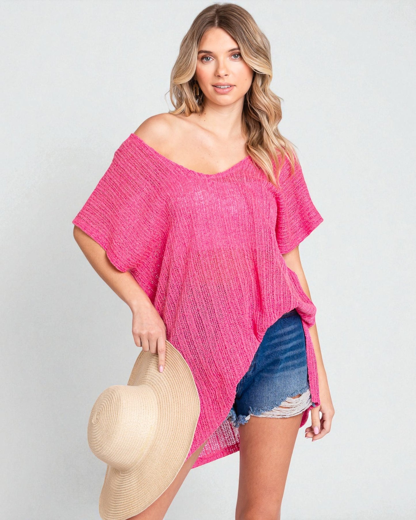 Woman wearing a pink off-shoulder top and denim shorts, holding a beige hat on a light gray background