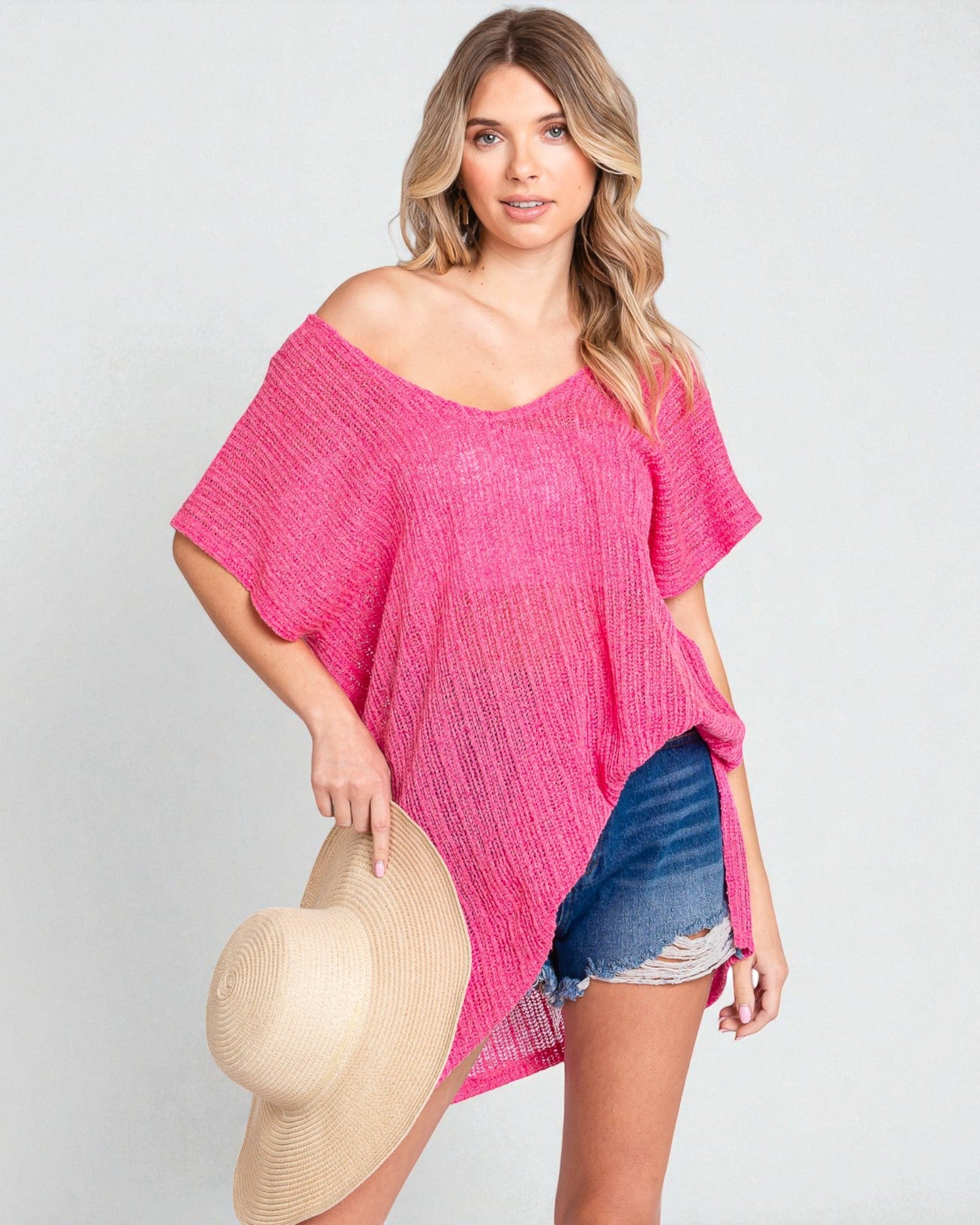 Woman wearing a pink off-shoulder top and denim shorts, holding a beige hat on a light gray background