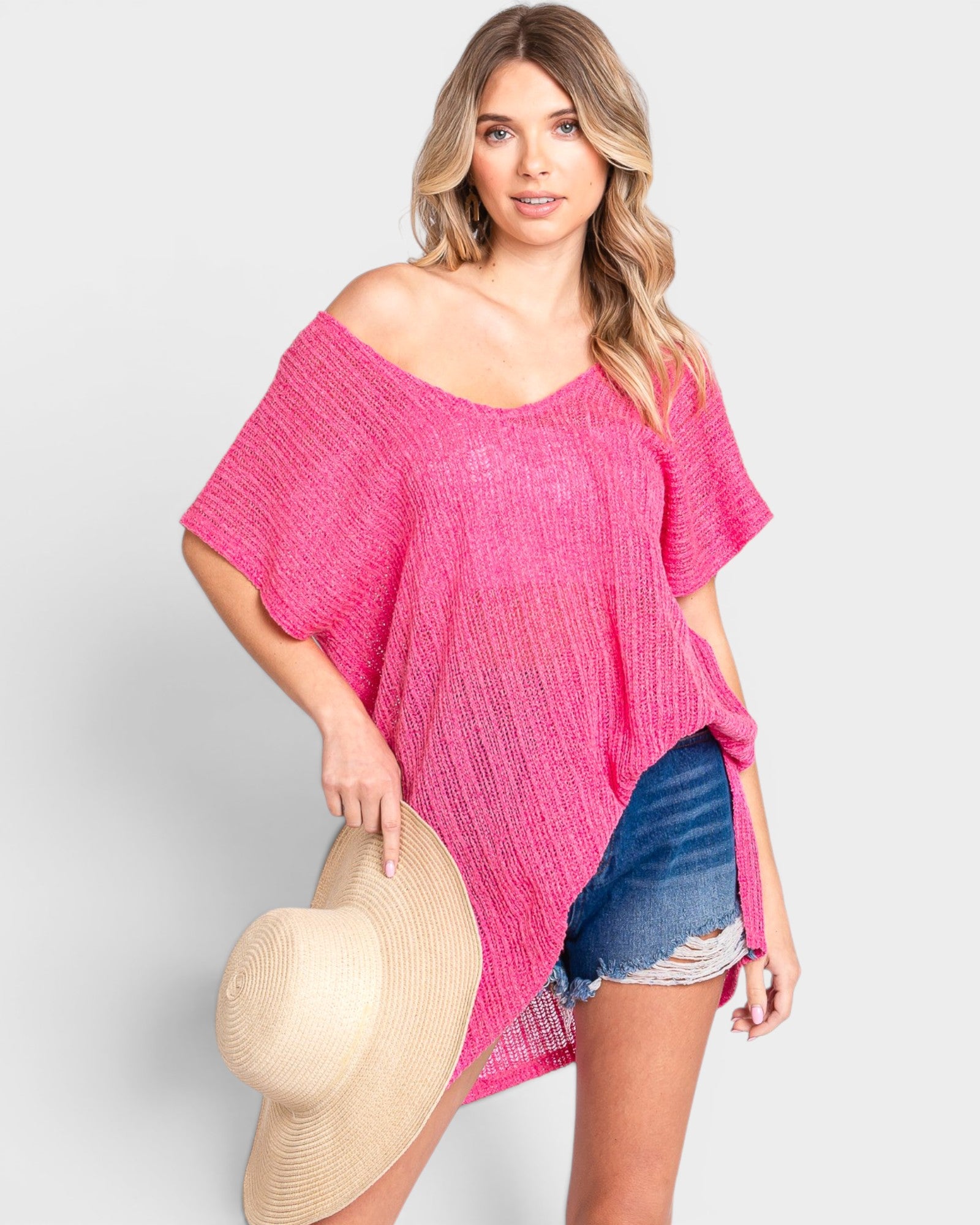 Woman wearing a pink off-shoulder top and denim shorts, holding a beige hat on a light gray background