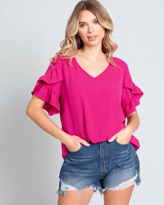 Woman wearing a bright pink blouse with ruffled sleeves and denim shorts on a gray background