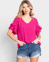 Woman wearing a bright pink blouse with ruffled sleeves and denim shorts on a light gray background
