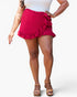 A woman wearing a bright pink wrap-style mini skirt with ruffle trim and a side tie detail, paired with a white top and tan platform sandals, against a light gray studio background.