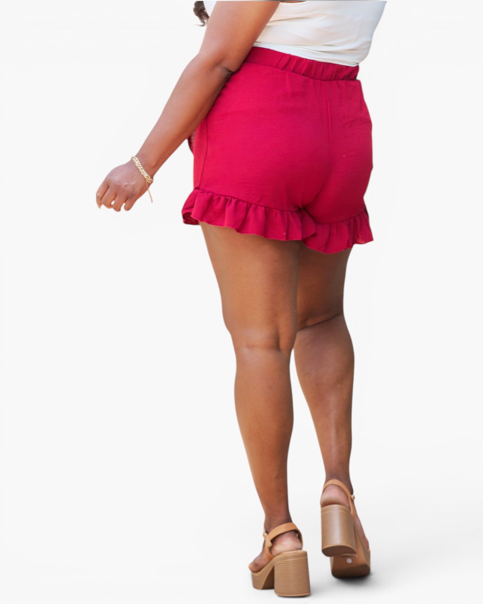 A person shown from the back wearing bright pink ruffled shorts, a white top, and tan platform sandals against a plain light gray studio background.