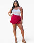 A woman modeling a white camisole top and a bright magenta ruffled mini skirt with side ties, paired with tan platform sandals, against a light gray studio background.