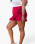A woman wearing a bright pink ruffled mini skirt with a side tie detail, paired with a white top and beige sandals, shown from the waist down against a light gray background.