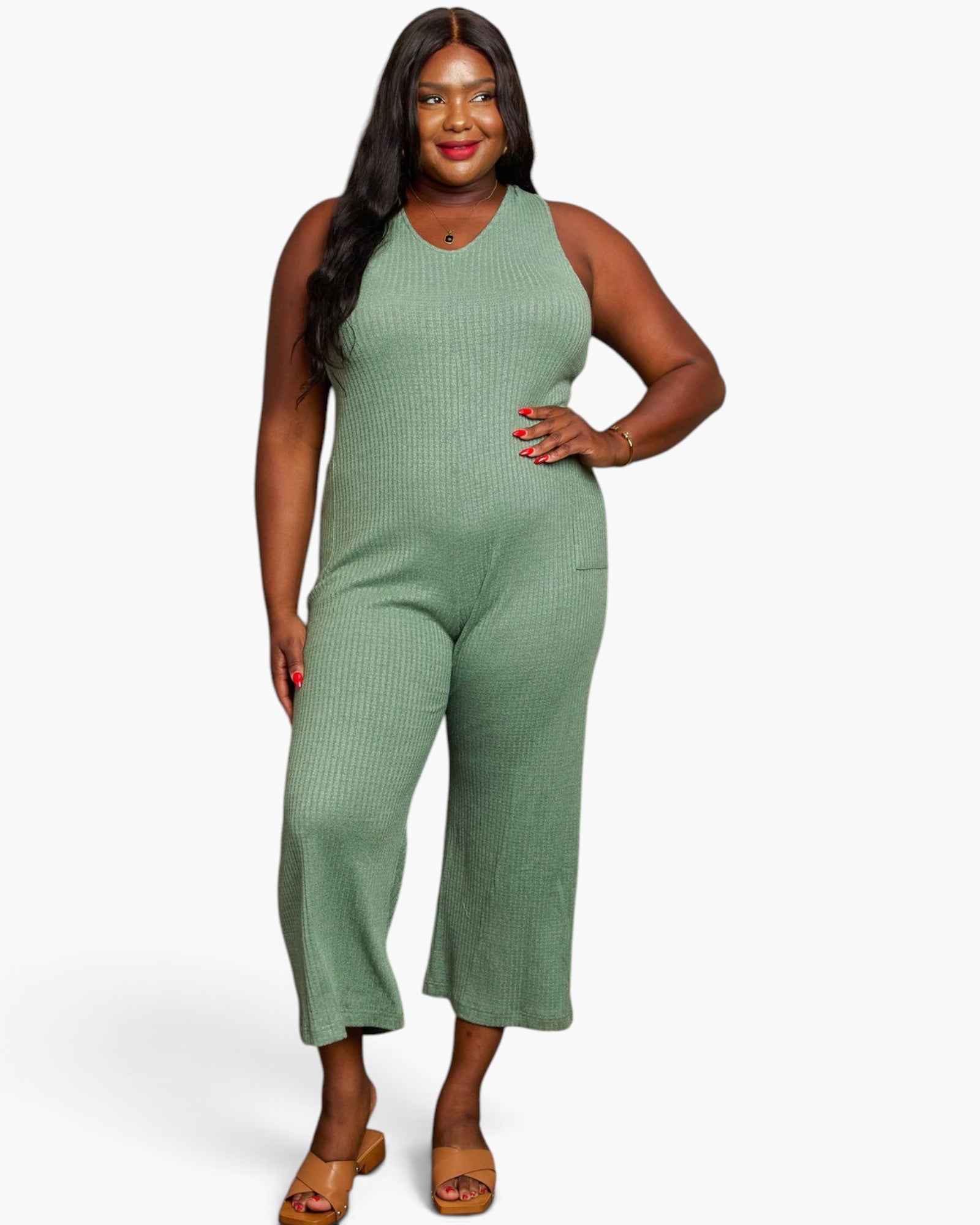 A woman modeling a sleeveless ribbed knit jumpsuit in sage green, paired with tan heeled sandals, against a plain light gray studio background.