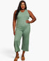 A woman modeling a sleeveless ribbed knit jumpsuit in sage green, paired with tan heeled sandals, against a plain light gray studio background.