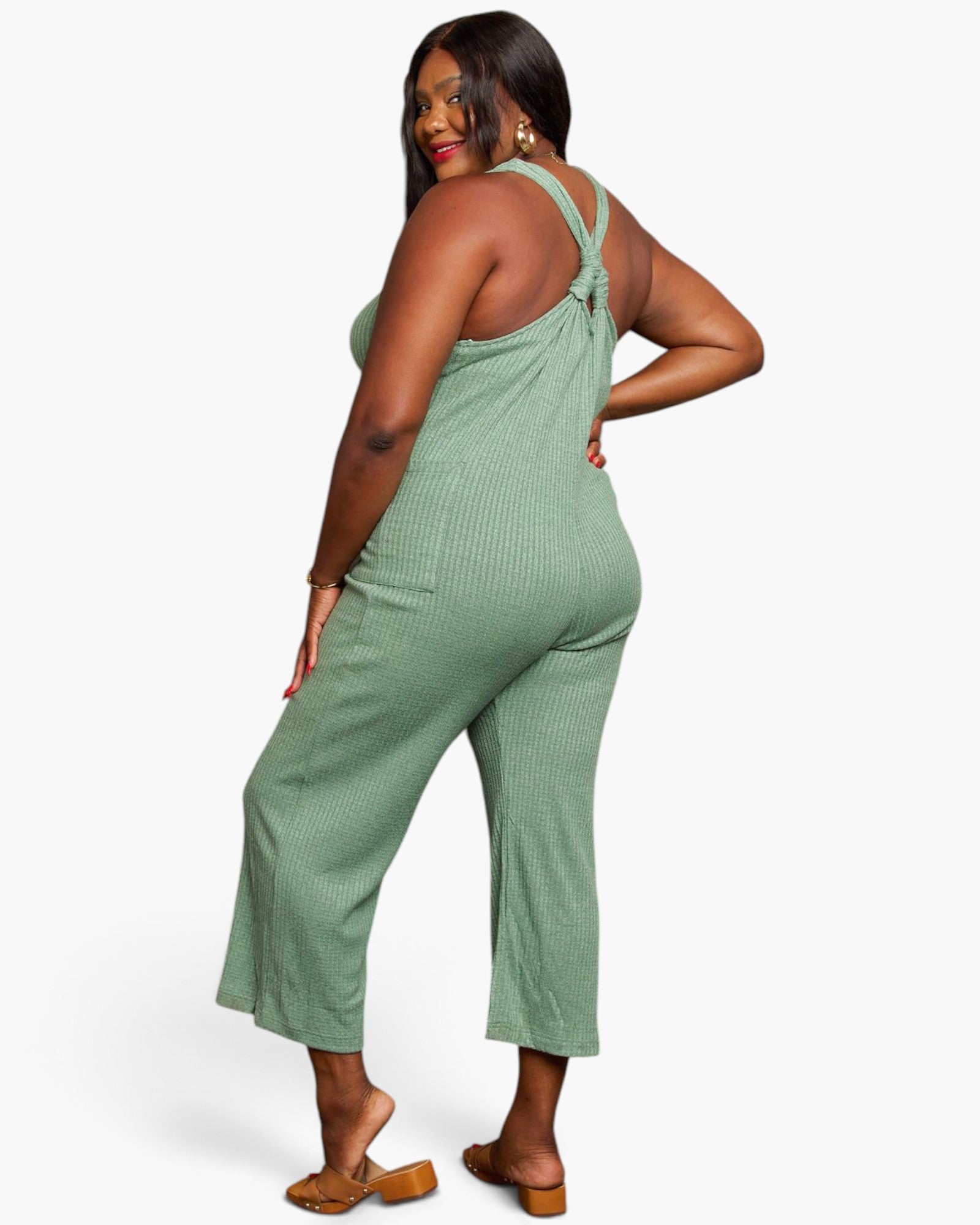 A woman modeling a sage green ribbed knit jumpsuit with a tie-shoulder back, shown from a three-quarters back view against a light gray studio background.