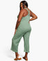 A woman modeling a sage green ribbed knit jumpsuit with a tie-shoulder back, shown from a three-quarters back view against a light gray studio background.
