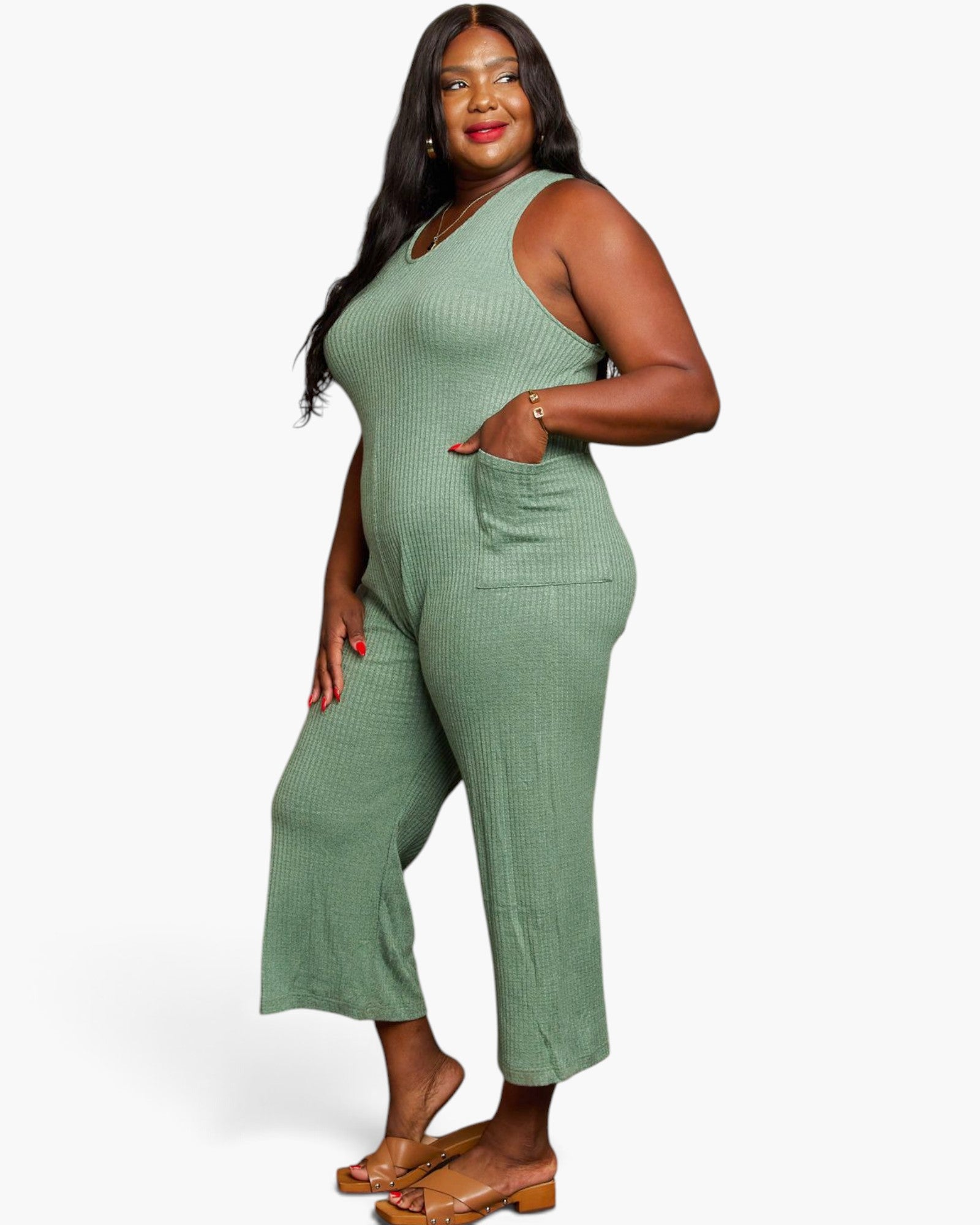 A woman modeling a sage green ribbed sleeveless jumpsuit with wide legs and a front pocket, paired with brown flat sandals, against a plain white studio background.