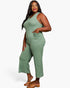 A woman modeling a sage green ribbed sleeveless jumpsuit with wide legs and a front pocket, paired with brown flat sandals, against a plain white studio background.