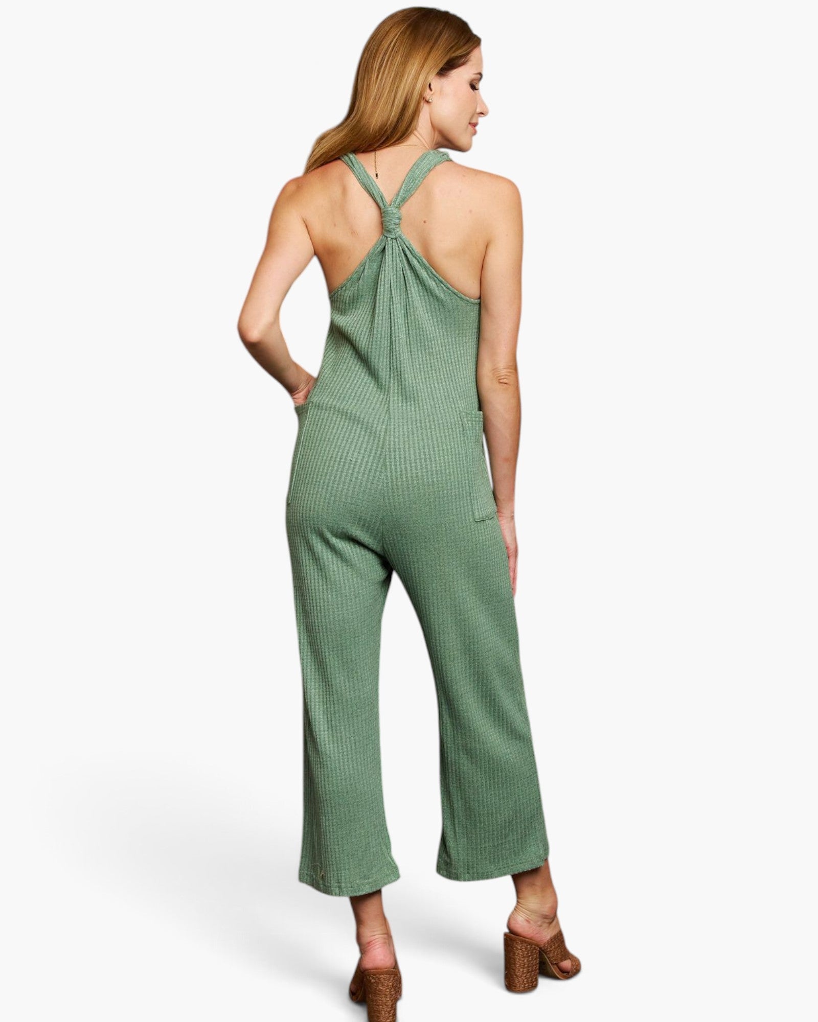A woman shown from the back wearing a sage green ribbed jumpsuit with a crossover halter-style back and wide cropped legs, paired with brown heeled sandals against a plain light background.