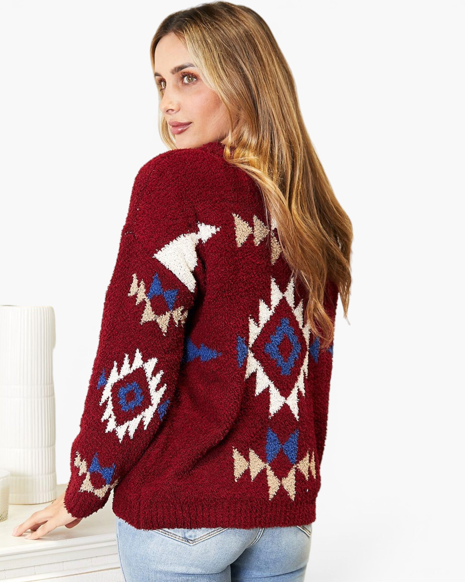A woman wearing a fuzzy knit cardigan with a Southwestern geometric pattern in burgundy, cream, and blue, paired with light blue jeans against a white background.