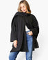 A woman wearing a black oversized wrap-style coat with a high draped collar, styled over a white top and blue jeans, against a plain light gray studio background.