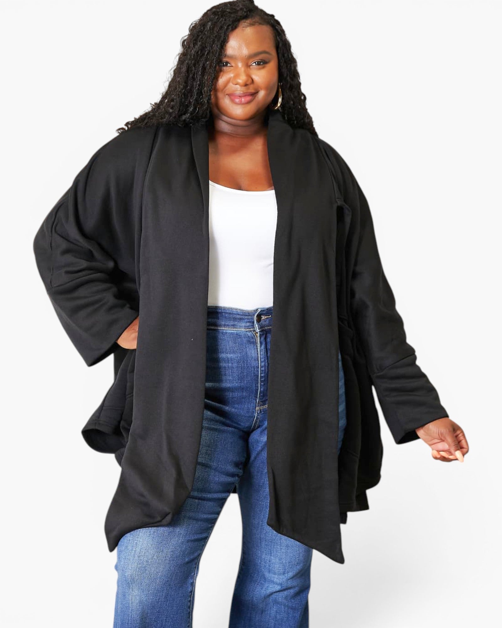 A woman modeling a black open-front wrap jacket over a white top and blue jeans, photographed against a plain light gray studio background.