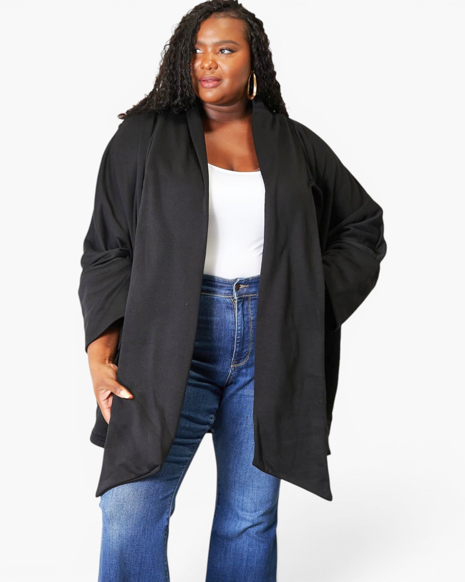 A woman modeling a black open-front cardigan or lightweight jacket over a white tank top and blue jeans, photographed against a plain light gray studio background.