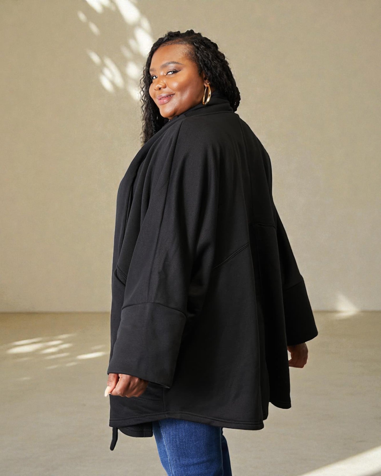 Woman wearing a black coat standing against a beige wall.