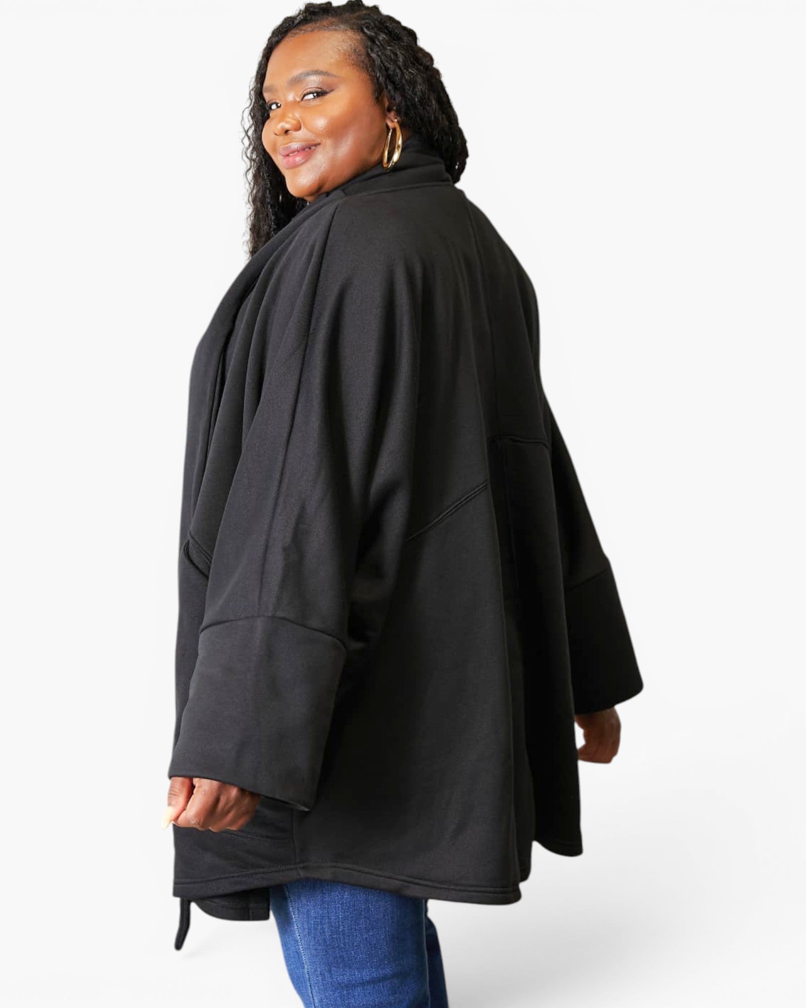 A woman wearing a loose black oversized jacket or cape-style top with wide sleeves, shown from the back and side against a light gray studio background.