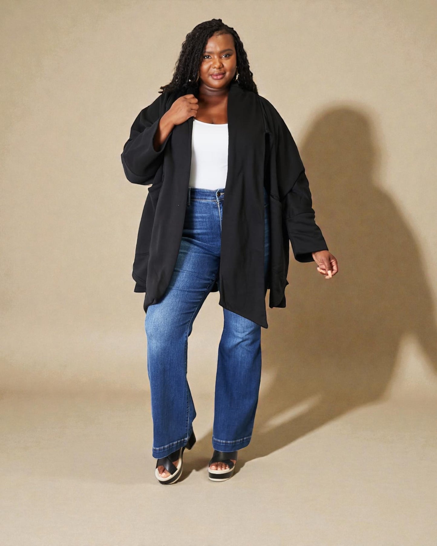 Woman wearing a black coat, white shirt, and blue jeans against a beige background