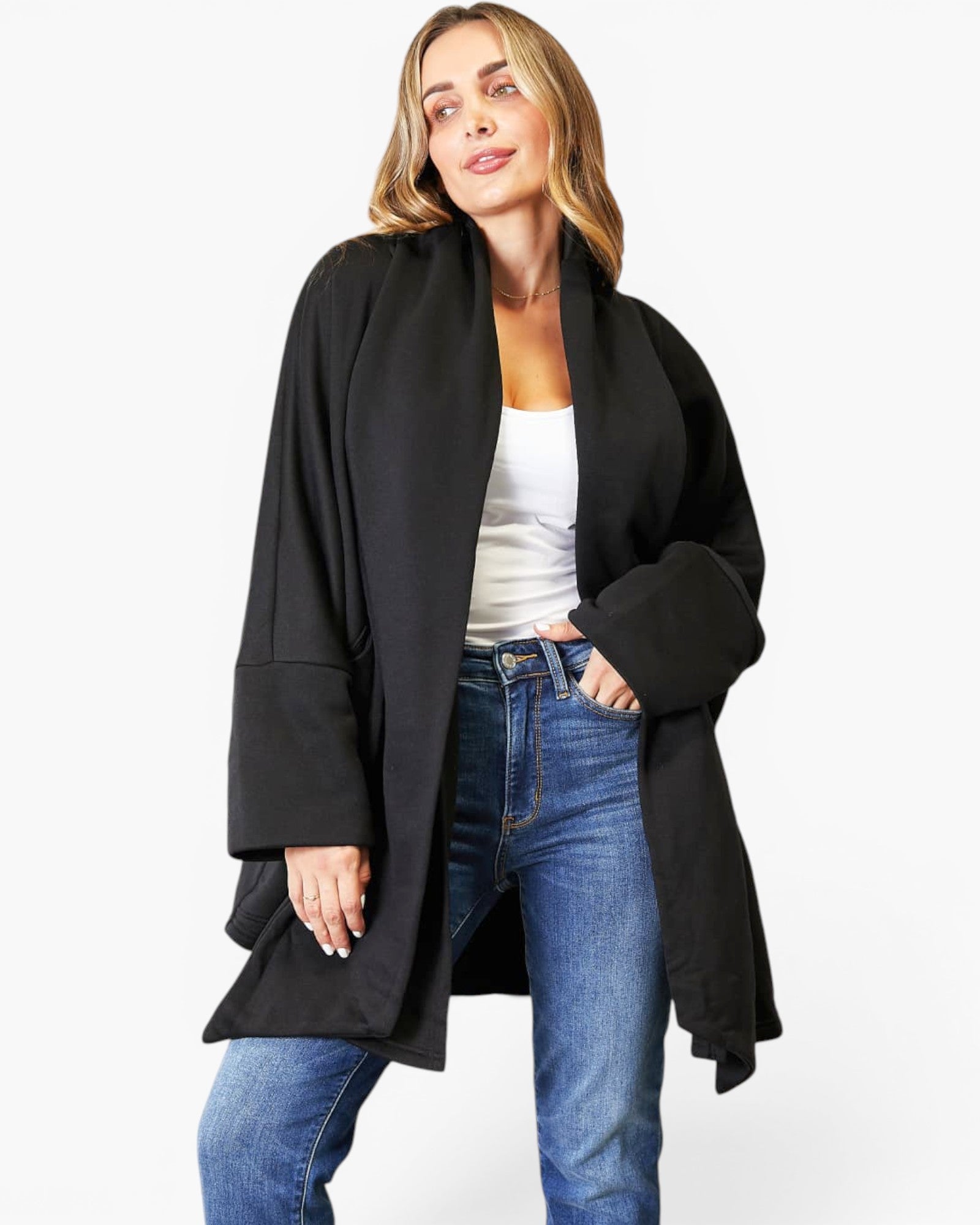 A woman modeling a black open-front shawl-collar coat with wide sleeves, worn over a white tank top and blue jeans, against a plain light gray studio background.