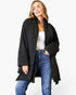 A woman modeling a black open-front shawl-collar coat with wide sleeves, worn over a white tank top and blue jeans, against a plain light gray studio background.