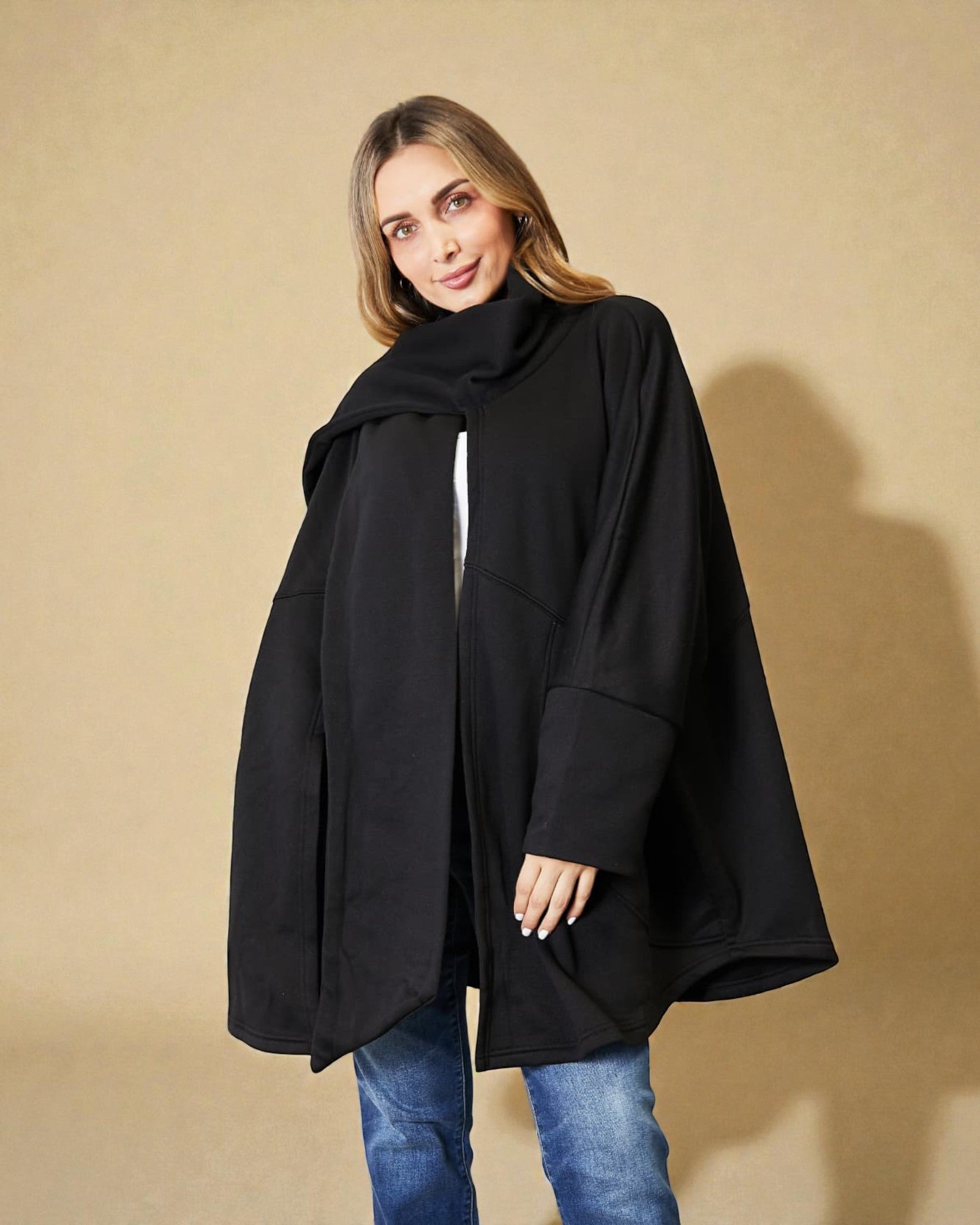 Woman wearing a black poncho over a white shirt with blue jeans on a beige background