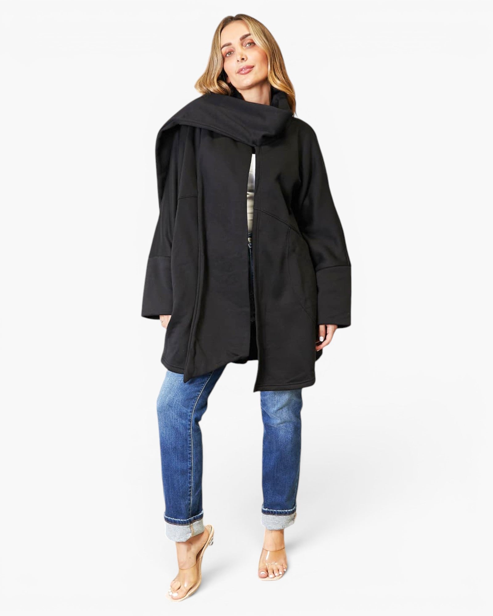 A woman modeling a black oversized wrap coat with a wide collar, worn over a patterned top and blue jeans, against a plain white studio background.