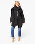 A woman modeling a black oversized wrap coat with a wide collar, worn over a patterned top and blue jeans, against a plain white studio background.