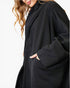 A close-up of a woman wearing an oversized black coat with wide sleeves and side pockets, shown against a plain white background.