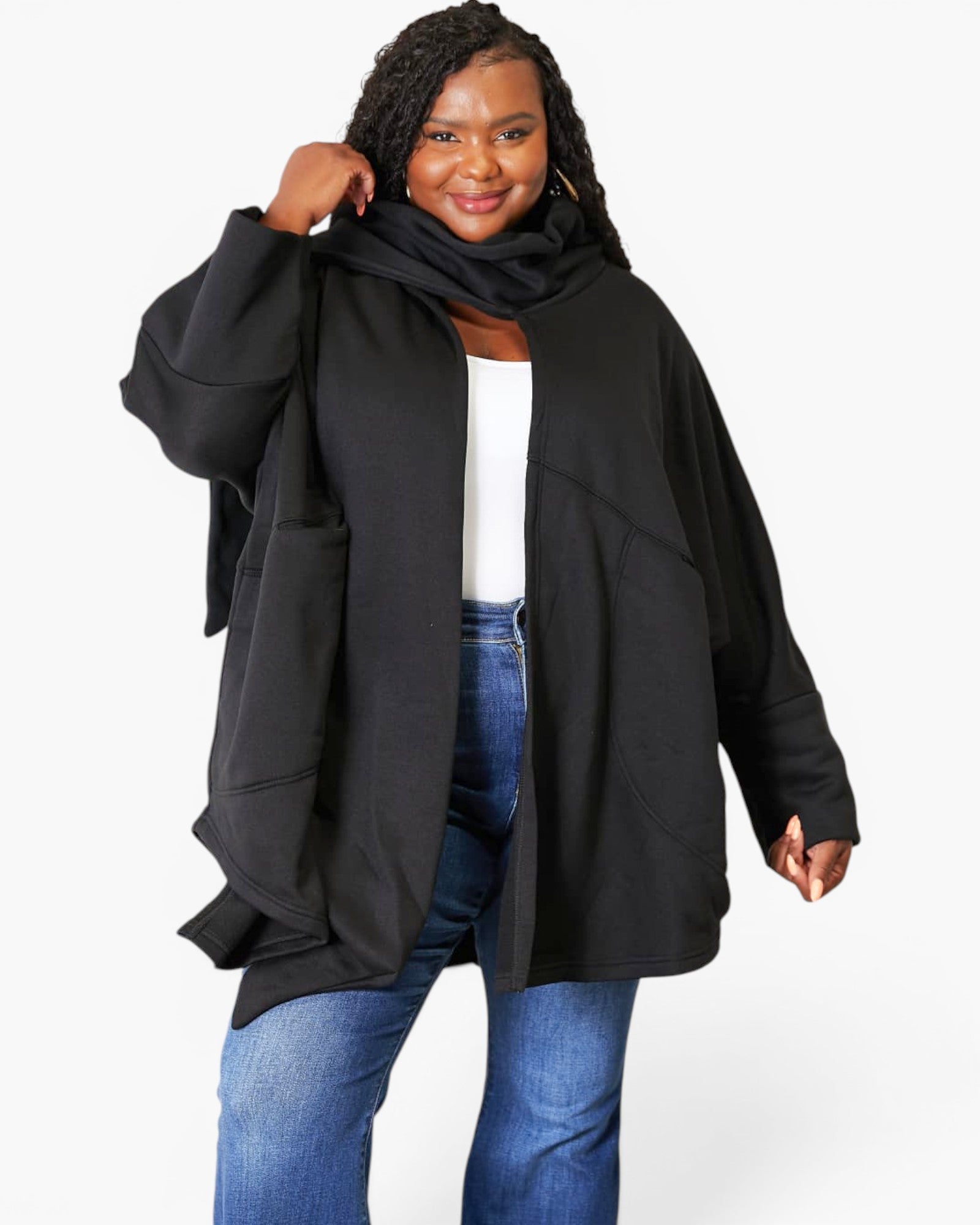 A woman modeling a black oversized open-front cardigan or jacket with a matching black scarf, worn over a white top and blue jeans, against a plain light gray studio background.