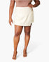 A model wearing a light beige wrap-style mini skirt, paired with a matching beige top and clear high-heeled sandals, against a plain white studio background.